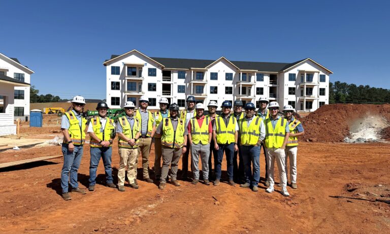 A group of people wearing safety vests and hard hats stand on a construction site in front of a partially built apartment building, representing the Columbus Office of Brasfield and Gorrie.