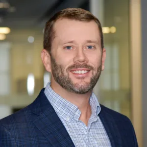 A man with short light brown hair and a beard, wearing a blue checked blazer and collared shirt, stands indoors in a modern office setting, smiling at the camera.