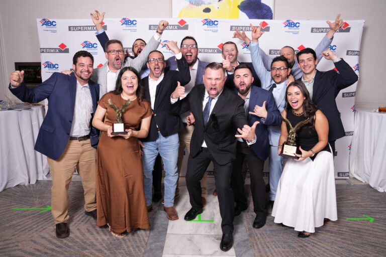 Un grupo de personas vestidas de gala posa con entusiasmo junto a unos trofeos delante de un telón de fondo con los logotipos de «SUPERMIX» y «ABC» en un evento celebrado en un recinto cerrado, representando con orgullo a la oficina de Miami de Brasfield and Gorrie.