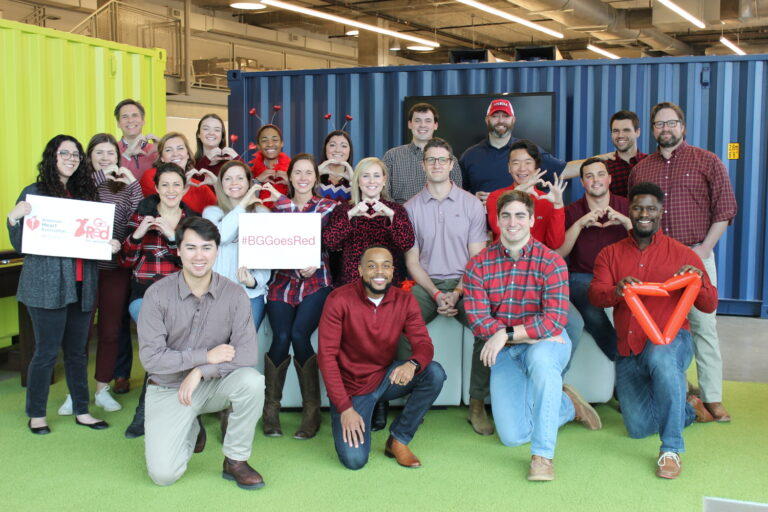 Grupo de personas sonrientes en un entorno de oficina informal, algunas con objetos en forma de corazón y vestidas de rojo, posiblemente apoyando una campaña de concienciación sobre la salud del corazón.
