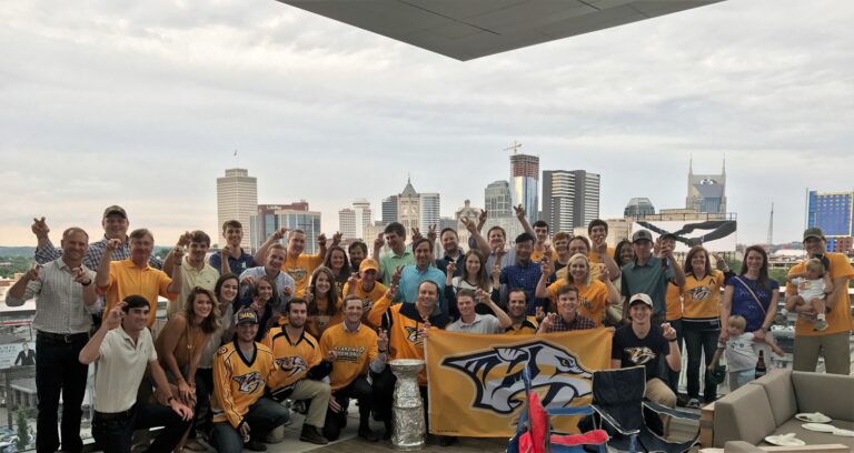 Un gran grupo de personas en una habitación con el horizonte de la ciudad de fondo, algunos con atuendos de equipos deportivos y posando para una foto de grupo.