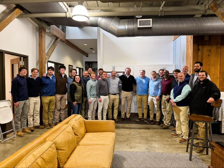 A group of around twenty men stand and pose for a group photo in the Columbus Office of Brasfield and Gorrie, set in a modern space with exposed beams and industrial vents.