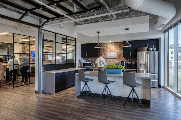 Modern Charlotte Office kitchen by Brasfield and Gorrie featuring island seating, a brick accent wall, stainless steel appliances, pendant lighting, and glass-walled meeting rooms in the background.