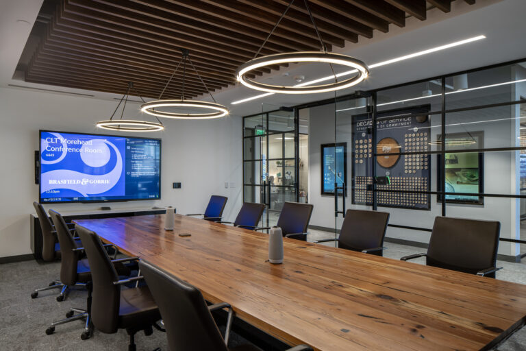 Modern conference room in the Charlotte office of Brasfield and Gorrie, featuring a long wooden table, black chairs, circular ceiling lights, a large wall-mounted screen, and glass walls displaying marketing materials.