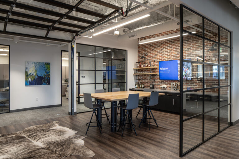 Una moderna sala de reuniones de Brasfield and Gorrie, con una mesa de madera y cuatro taburetes, una pared acristalada y un televisor que muestra un mensaje de «Bienvenida» sobre una pared de ladrillo en la oficina de Charlotte.