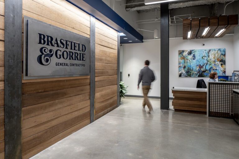 A person walks through the Charlotte Office of Brasfield & Gorrie General Contractors, featuring wood panel walls, modern lighting, and a reception desk with a receptionist.