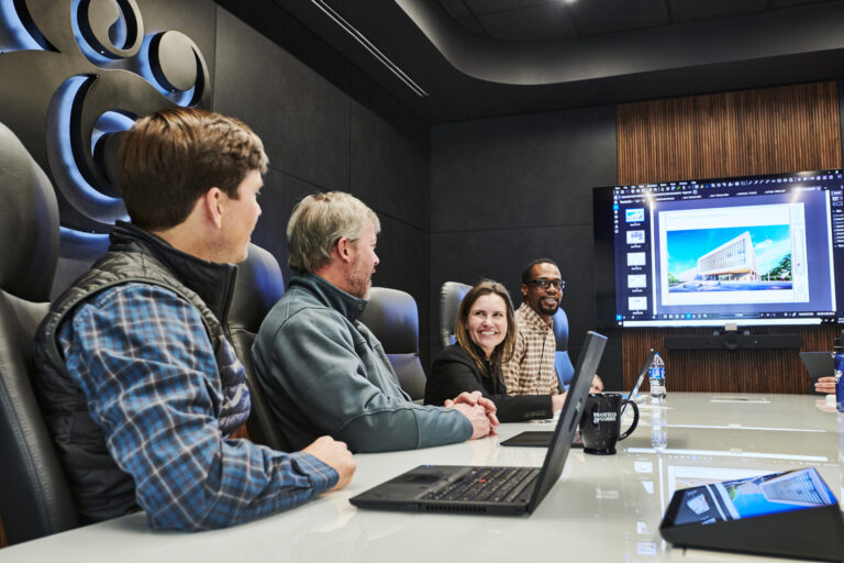 Four professionals in a modern conference room engaging in a collaborative discussion with a presentation on a screen.