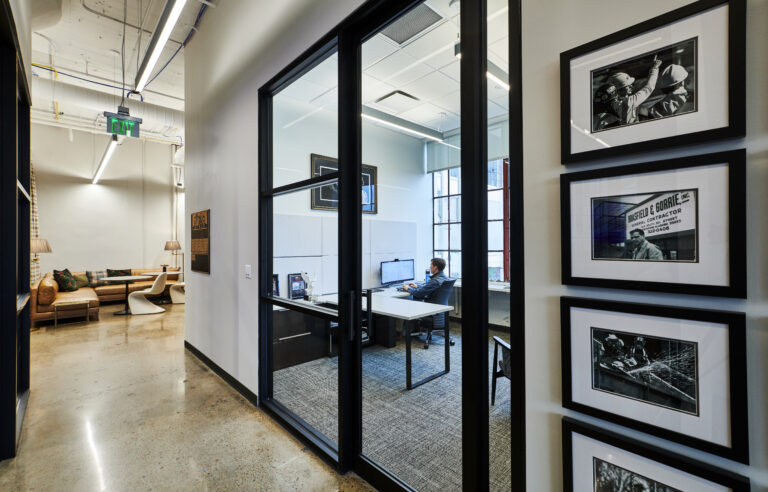 Modern office space with framed artwork on the walls and an individual working at a computer desk visible through a glass wall.