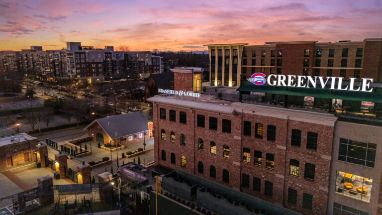 Aerial view of downtown Greenville, SC at sunset, highlighting offices, apartments, and the large illuminated Greenville sign on a brick building near the Greenville Office Brasfield and Gorrie.