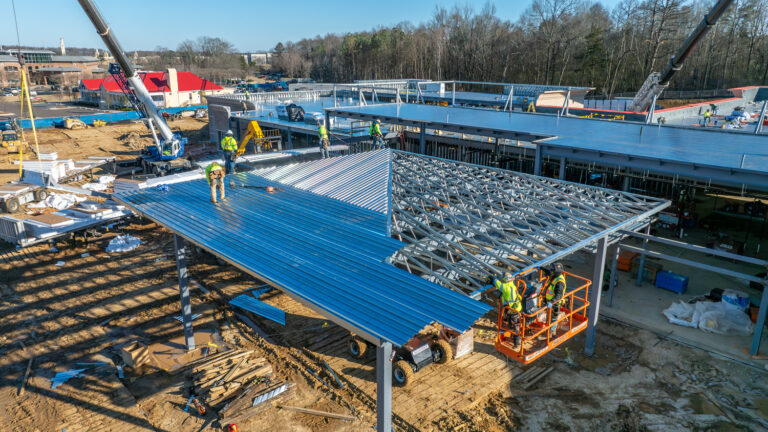 Construction workers install large blue metal roof panels on the Jackson Office building structure at a construction site, with cranes and equipment from Brasfield and Gorrie visible around the area.