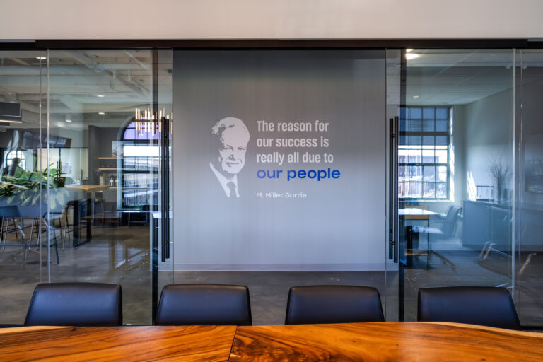 A glass-walled conference room in the Greenville Office features a wall graphic of a man’s face and the quote, “The reason for our success is really all due to our people – M. Miller Gorrie.”.