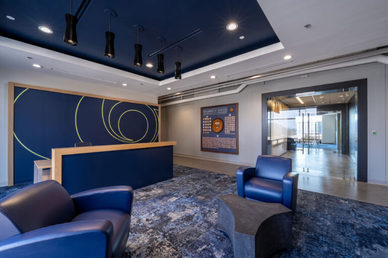 Modern office lobby in the Greenville Office Brasfield and Gorrie building with blue chairs, abstract wall art, a wooden reception desk, and a glass door leading to a hallway with windows.