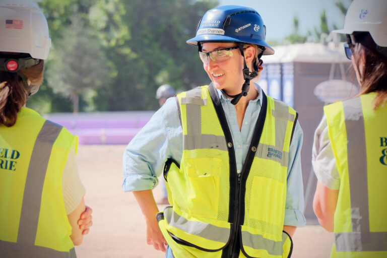 At a construction site, an Orlando Office Brasfield and Gorrie team member in a hard hat, safety goggles, and yellow vest talks with two others wearing similar protective gear.