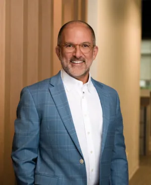A man with glasses and a beard wearing a blue plaid suit jacket and white shirt stands indoors by a wooden wall, smiling at the camera.