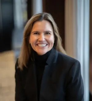 Sara Beth Wilcox, a woman with shoulder-length light brown hair, smiles indoors while wearing a black blazer and black turtleneck.