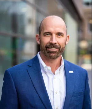 Ben Barfield, a bald man with a beard, stands outside near glass buildings in a blue suit jacket and white dress shirt, looking at the camera and smiling.