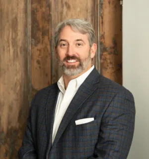Wes Kelley, LEED AP BD+C, un hombre de cabello canoso y barba que lleva una chaqueta de traje a cuadros azules y una camisa blanca, posa delante de un fondo rústico de madera.