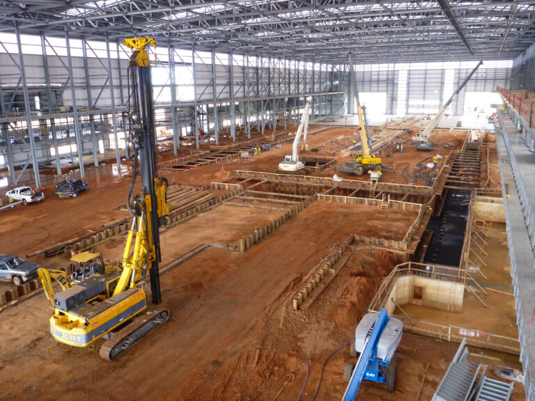 Gran obra de construcción cubierta con múltiples excavadoras y maquinaria trabajando en amplias excavaciones de infraestructuras bajo un armazón metálico.