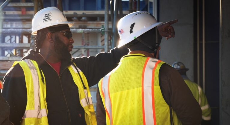 Two construction workers in high-visibility vests and helmets discussing their careers on a building site.