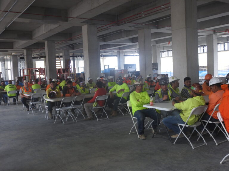 Construction workers in safety gear celebrating over lunch at tables inside a partially completed building