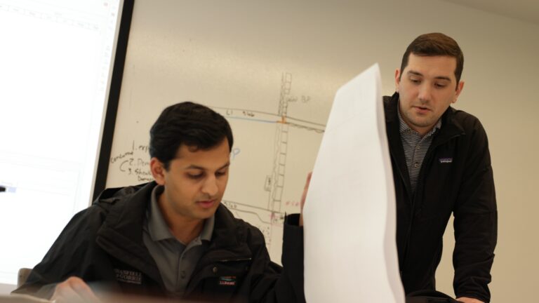 Two men examining a large paper chart related to their careers in an office with a whiteboard featuring technical drawings in the background.