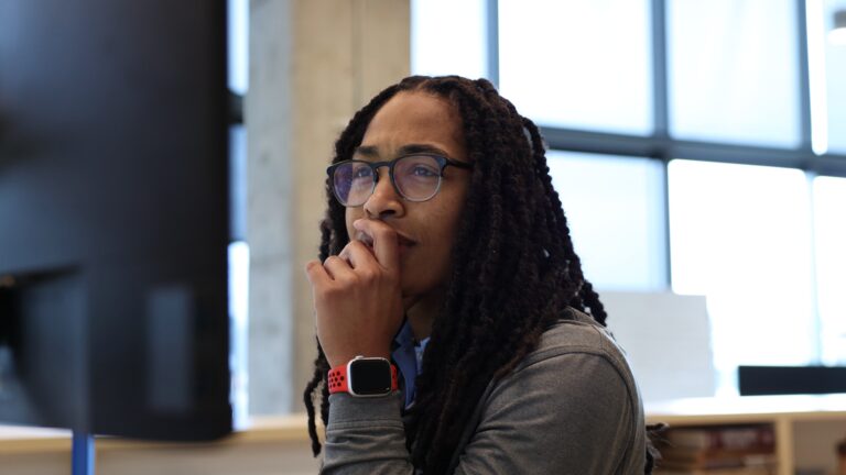 A thoughtful woman with glasses and braided hair, wearing a smartwatch, is resting her chin on her hand while looking at a desktop monitor in an office environment.