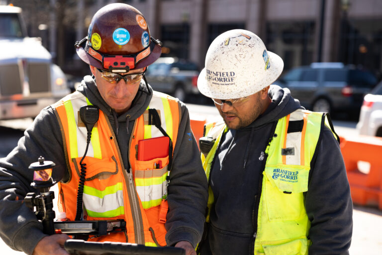 Two construction workers in high visibility jackets and hard hats examine a digital tablet at a worksite.