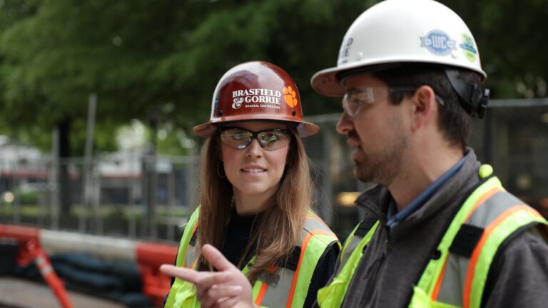 A female and male construction worker in hard hats and safety vests having a discussion at a construction site.