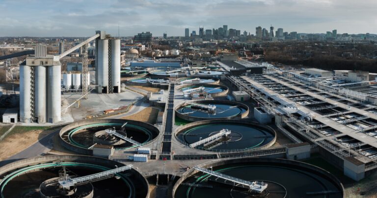 Aerial view of an urban wastewater treatment plant with multiple circular settling tanks, concrete structures, and industrial buildings, all expertly constructed by Brasfield & Gorrie, with a city skyline visible in the background.