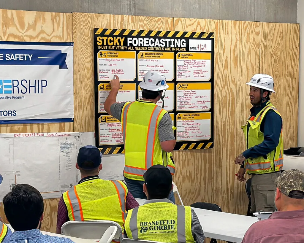 Two construction workers in hard hats and reflective vests engaging in a discussion in front of an “Auto Draft” board in a site office.
