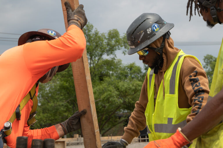 Three construction workers in safety gear conversing around a wooden pole at a job site.