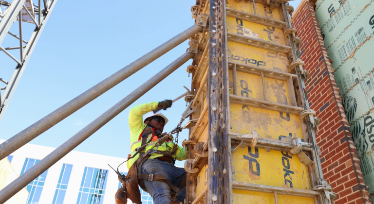 Construction worker in a high-visibility jacket securing a safety harness while climbing a yellow scaffolding structure.