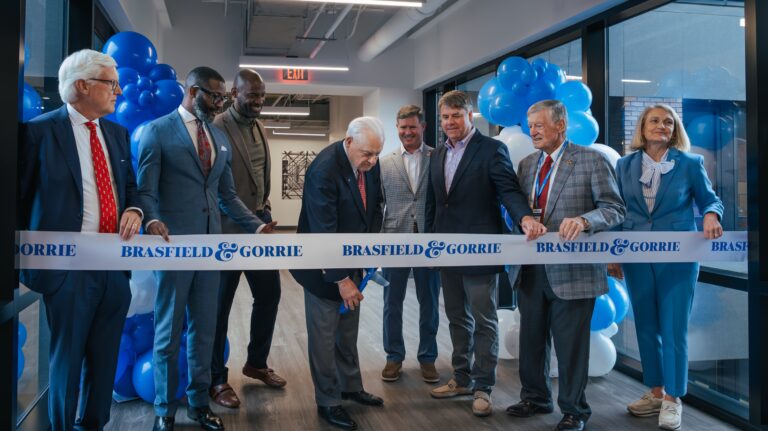 A group of people stand behind a ribbon labeled “Brasfield & Gorrie” as one man prepares to cut it at an indoor ribbon-cutting ceremony with blue and white balloons, celebrating the company's headquarters expansion.