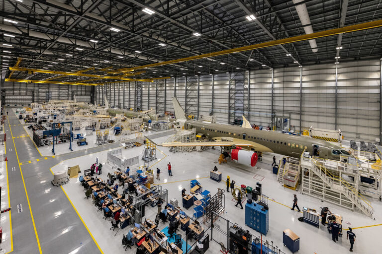 A large aircraft assembly plant with partially built airplanes, workers, workstations, and machinery under bright overhead lighting showcases self perform construction in a spacious industrial facility.