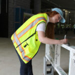 A person wearing a yellow safety vest and blue cap writes on a white beam inside a construction site.