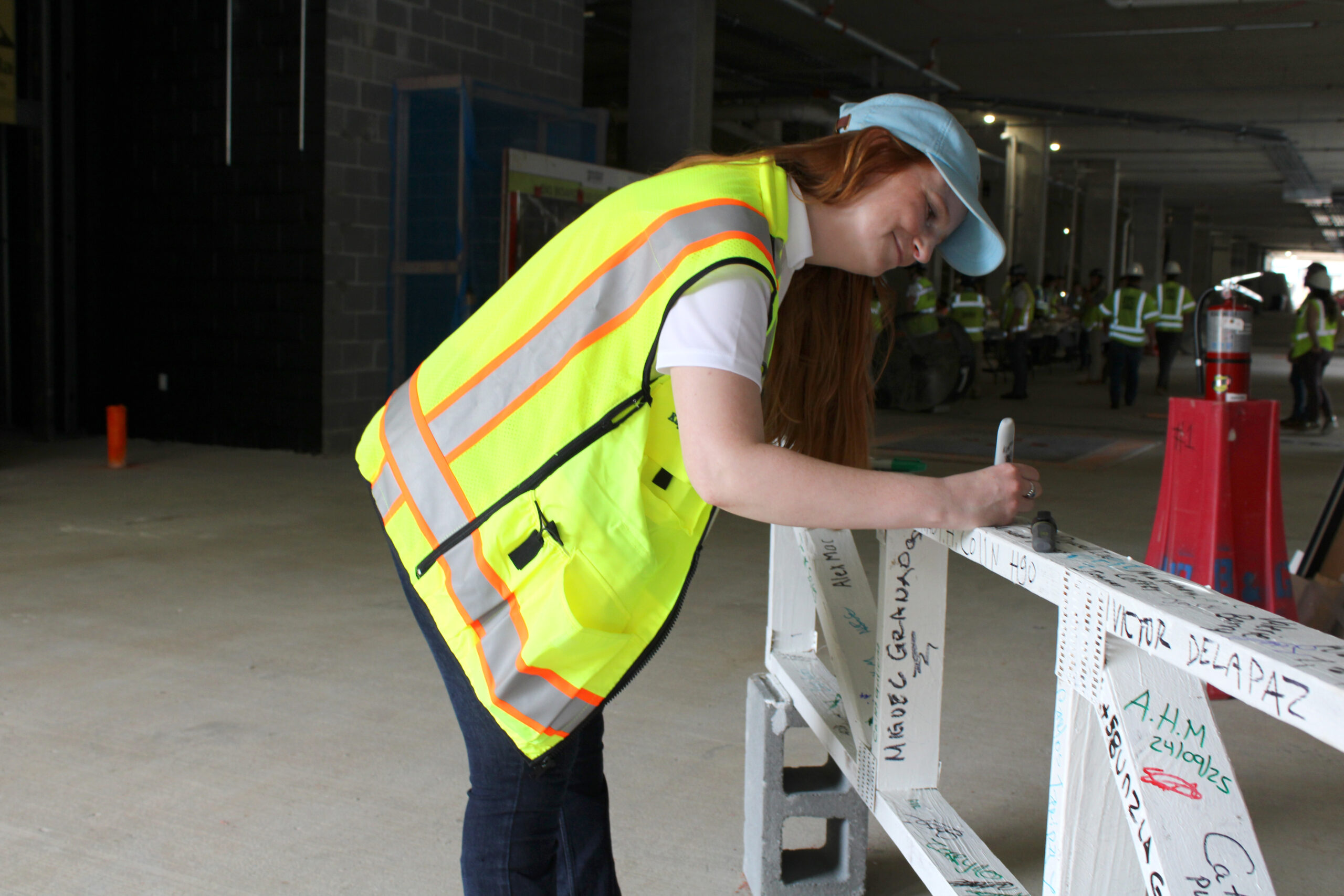 A person wearing a yellow safety vest and blue cap writes on a white beam inside a construction site.