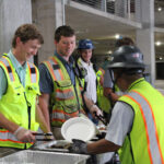 Construction workers in safety vests and helmets serve food onto plates at a buffet-style table in an under-construction setting.