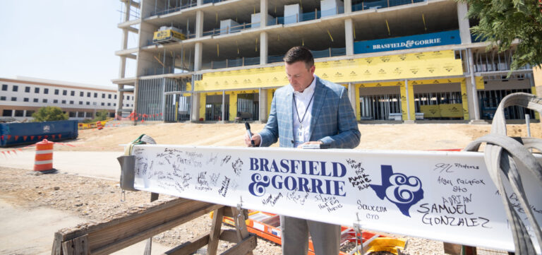 A man signs a white steel beam covered in signatures at a Central Texas construction site, with a partially completed multi-story building rising in the background.