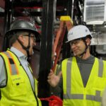 Two people wearing hard hats and safety vests at an industrial worksite with metal structures and a ladder in the background.