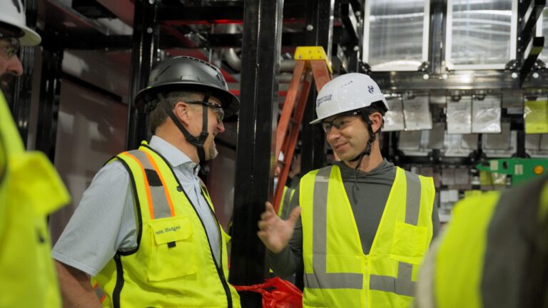 Two people wearing hard hats and safety vests at an industrial worksite with metal structures and a ladder in the background.