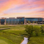 A golf course green with a flag in the foreground, a small creek, and a large modern building under a colorful Texas sunset sky in the background.