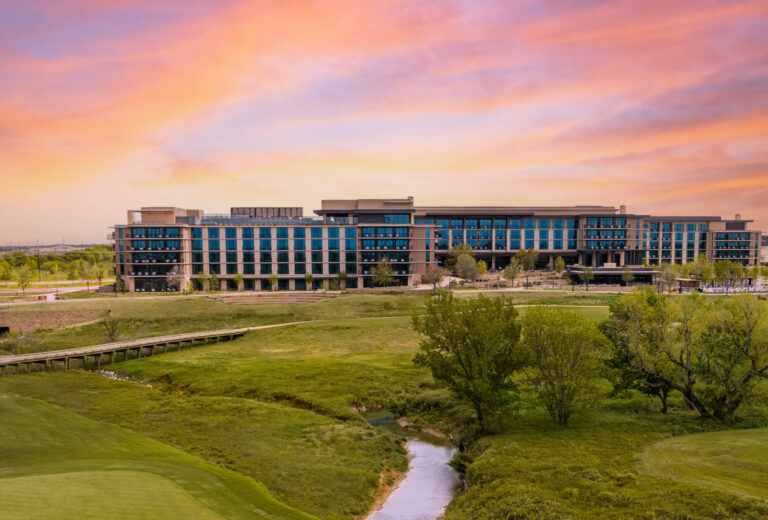 A golf course green with a flag in the foreground, a small creek, and a large modern building under a colorful Texas sunset sky in the background.