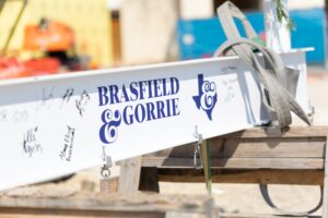 A white steel beam with "Brasfield & Gorrie" and a Texas outline logo, signed by various people, rests on wooden supports at a construction site.