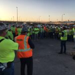 A large group of construction workers wearing safety vests and hard hats gather outdoors at a Texas job site during sunrise.