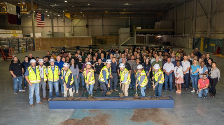A large group of people, some in construction vests and helmets, stand inside a spacious warehouse for the Airbus helicopter expansion groundbreaking ceremony with Brasfield & Gorrie.