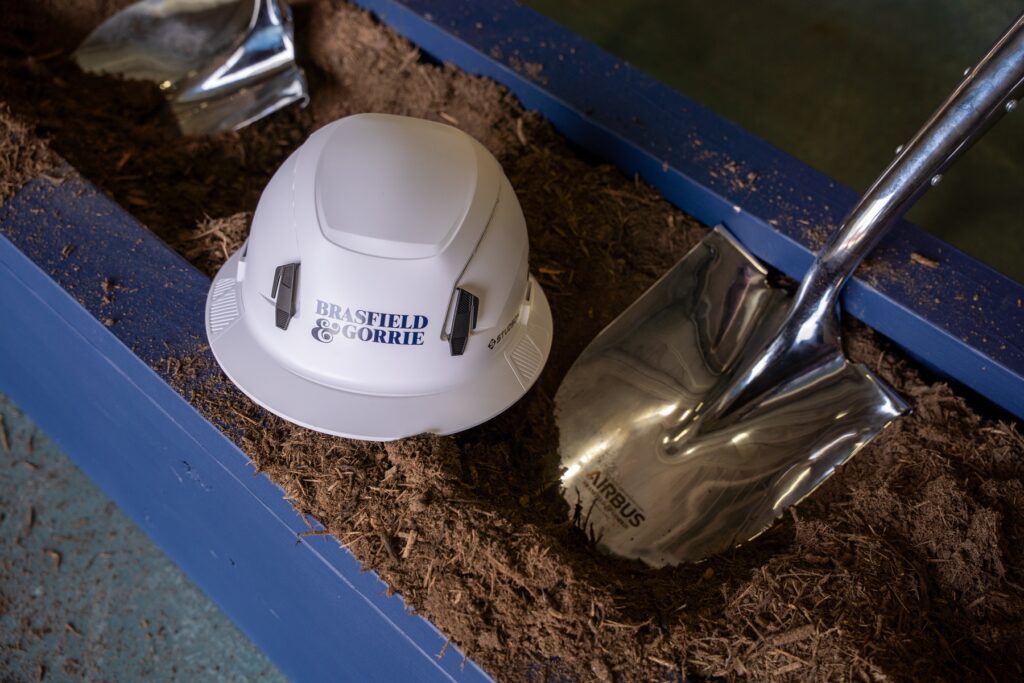 A white hard hat labeled "Brasfield & Gorrie" and a shiny metal shovel rest on soil inside a blue rectangular container, symbolizing the groundbreaking of the Airbus helicopter expansion construction project.