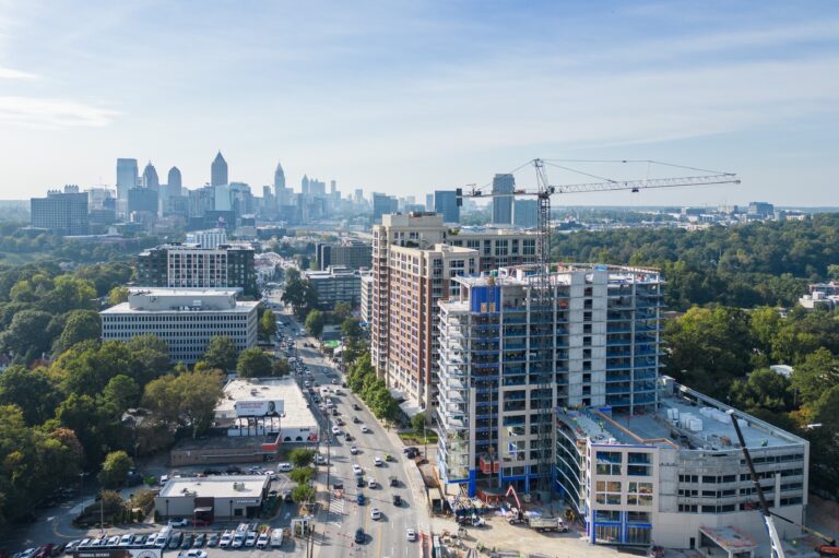Aerial view of a city with a high-rise building under construction by Brasfield & Gorrie, a crane at work, busy streets below, and a skyline of tall buildings in the distance.