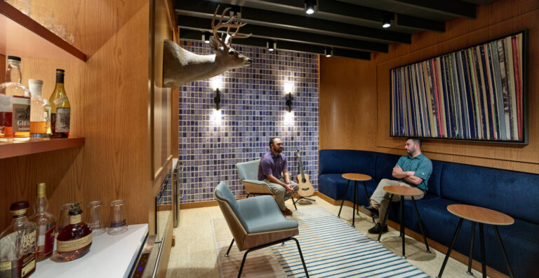 Two people sit and talk in a modern lounge with wood-paneled walls, a mounted deer head, a guitar, and shelves with liquor bottles.