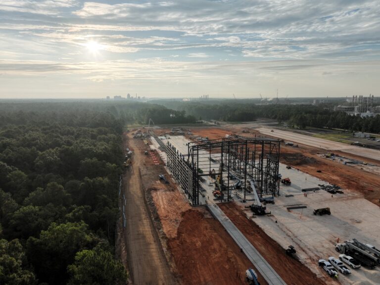 Vista aérea de una obra de Brasfield & Gorrie con armazón de acero que se eleva sobre una gran cimentación de hormigón, bordeada de tierra roja y zonas boscosas bajo un cielo parcialmente nublado.