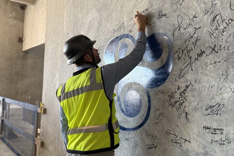 A person in a safety vest and hard hat signs a concrete wall covered with signatures at the future Westchester emergency care and specialty care facility construction site.